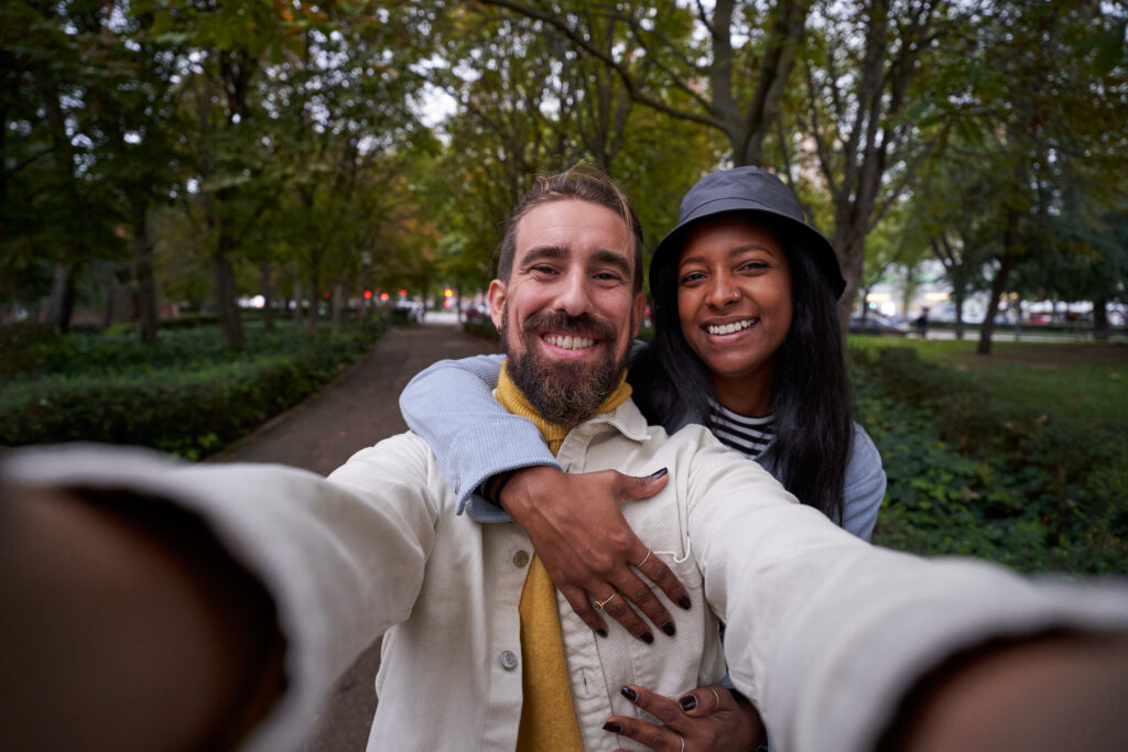 Cheerful selfie of young multiethnic couple looking at camera smiling, outdoors in park. Copy space.