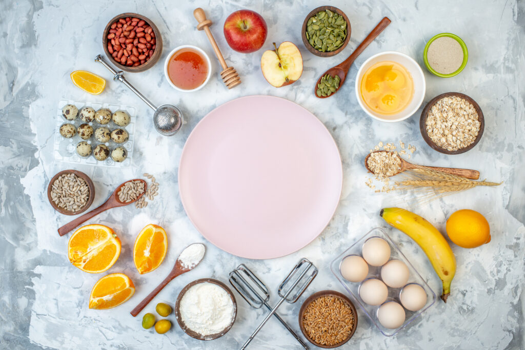 A top view of a plate on a table surrounded by different types of nutrition, such as: fruits, eggs, seeds, and vegetables.